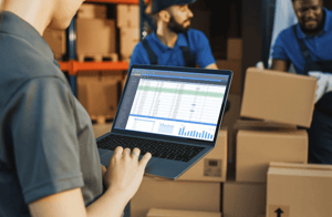 Person checking numbers on a laptop while workers move boxes in the background