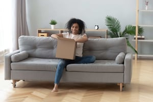 Woman smiling as she opens a box while seated on her couch-min